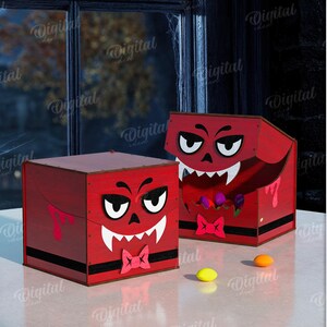 K&ouml;nnte beinhalten: Zwei rote Halloween-Boxen mit Cartoon-Teufelsgesichtern, wei&szlig;en Fangz&auml;hnen und rosa Fliegen. Die Boxen stehen auf einer wei&szlig;en Oberfl&auml;che mit einem K&uuml;rbis und einem Fenster im Hintergrund. Die Materialbreite betr&auml;gt 3 mm.