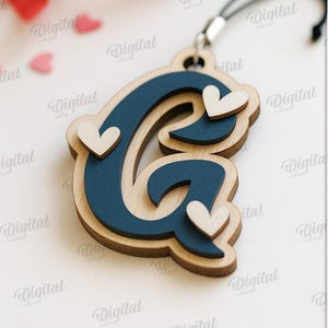 May include: Wooden layered letter "G" pendant in navy blue and natural wood tones, adorned with three small white hearts. The pendant hangs from a black cord. The image also includes the text "SUITED FOR ANY MATERIAL THICKNESS".