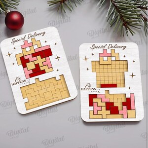 May include: Two wooden puzzle pieces with a Tetris-like design in red, pink, and tan colors. The words "Special Delivery" and "Fit Happens" are printed on the pieces. A red Christmas ornament and pine branches are in the background.