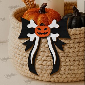 K&ouml;nnte beinhalten: Eine dekorative Halloween-Schleife mit einem geschnitzten K&uuml;rbisgesicht und Knochenakzenten. Die Schleife ist schwarz und wei&szlig;, mit einem geschnitzten K&uuml;rbis in der Mitte. Sie befindet sich in einem geflochtenen Korb mit K&uuml;rbissen und Kerzen.