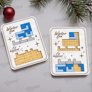 May include: Two wooden puzzles with the text "Winter fun" and "Fit happens". The puzzles feature a blue and gray design, with a light brown base. A red ornament and evergreen branches are in the background.