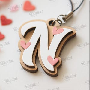 May include: Wooden letter "N" pendant with a white top layer and brown base, adorned with pink hearts. The pendant is attached to a black cord. The image also includes scattered red heart shapes and the text "SUITED FOR ANY MATERIAL THICKNESS".
