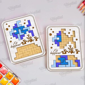 May include: Two wooden puzzle boards with blue and light blue pixelated designs, brown coral accents, and the text "Fit happens." The boards are rectangular with rounded corners. The image also includes a watercolor palette and colored pencils.