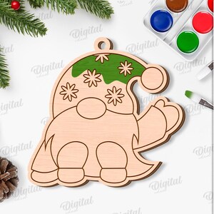 May include: A wooden gnome-shaped Christmas ornament with a green hat decorated with white stars. The ornament is unfinished and ready for painting. The image also includes paint, brushes, and text that says "SUITED FOR ANY MATERIAL THICKNESS".