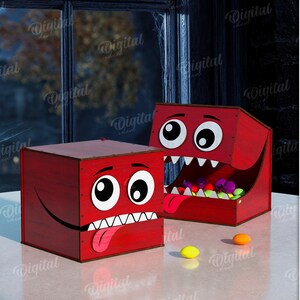 K&ouml;nnte beinhalten: Zwei rote, w&uuml;rfelf&ouml;rmige Boxen mit Cartoon-Monstergesichtern. Eine Box hat einen offenen Mund voller bunter B&auml;lle. Die andere Box hat einen geschlossenen Mund, aus dem eine Zunge herausragt. Die Boxen stehen auf einem Tisch.