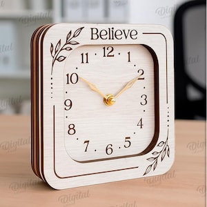 K&ouml;nnte beinhalten: Eine quadratische Holzuhr mit dem Wort &bdquo;Believe&ldquo; oben. Das Zifferblatt hat schwarze Ziffern und goldfarbene Zeiger. Dekorative Blattmotive befinden sich an den Seiten. Die Uhr ist 3 mm breit.