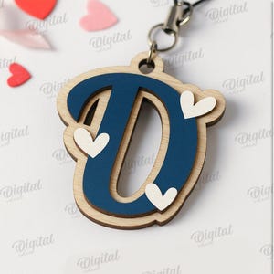 May include: A wooden letter "D" pendant with a navy blue center and light wood border, adorned with three white heart accents. The pendant hangs from a silver-colored chain. Scattered around are red and pink heart-shaped decorations.