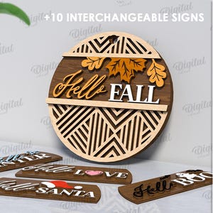 Könnte beinhalten: Ein rundes Holzschild mit den Worten "Hello FALL" und austauschbaren Schildern für verschiedene Jahreszeiten. Das Schild hat ein geometrisches Design und Herbstblatt-Akzente. Der Text "+10 INTERCHANGEABLE SIGNS" ist ebenfalls enthalten.