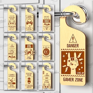 Puede incluir: Diez diseños diferentes para un colgador de puerta para jugadores. El colgador de puerta está hecho de madera y tiene un esquema de color marrón y negro. Los diseños incluyen frases como "Gaming in Progress", "No molestar" y "Gamer Zone".
