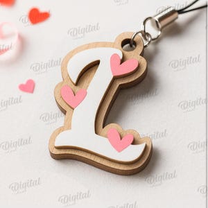 May include: Wooden letter "L" charm with a white overlay and pink heart accents. The charm is attached to a silver-colored lanyard. Scattered around are pink and red heart-shaped confetti. The text "SUITED FOR ANY MATERIAL THICKNESS" is visible.