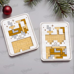 May include: Two wooden puzzle pieces with a light wood finish, featuring geometric shapes in gold and gray. The phrase "Fit Happens" is printed on each piece. A red ornament and evergreen branches are in the background.