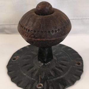 Pode incluir: Um puxador de porta antigo com um topo arredondado castanho escuro e texturizado e uma base circular preta decorativa. O puxador tem um topo pequeno e redondo e uma série de pequenos orifícios ao meio. A base tem um desenho floral.