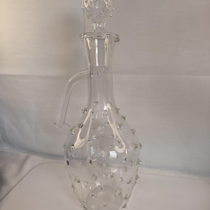 Könnte beinhalten: Transparente Glaskaraffe mit Griff und Verschluss. Die Karaffe hat einen bauchigen Körper und ist mit kleinen, klaren, blasenartigen Vorsprüngen verziert. Der Verschluss ist ebenfalls transparent und hat ein ähnliches Blasendesign. Die Karaffe ist zur Aufnahme von Flüssigkeiten bestimmt.