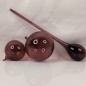 May include: Three glass ornaments in shades of purple with air bubbles inside. The ornaments are round and teardrop shaped.