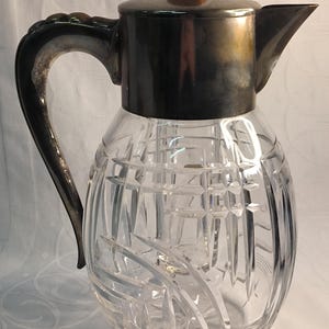 Könnte beinhalten: Eine klare, geschliffene Glaskaraffe mit einem silberfarbenen Metalldeckel und Griff. Der Deckel hat einen kleinen, runden, braunen Knopf. Der Körper der Karaffe weist ein geometrisches Muster auf.