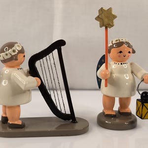 Könnte beinhalten: Zwei kleine hölzerne Engelsfiguren in weißen Gewändern mit dunkelblauen Flügeln, die mit goldenen Sternen verziert sind. Ein Engel spielt eine schwarze Harfe, der andere hält einen goldenen Sternenstab und eine gelbe Laterne auf grauen Sockeln.