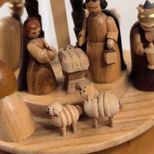 Könnte beinhalten: Eine hölzerne Krippenszene mit geschnitzten Figuren einer Person mit langem dunklem Haar, einer Person mit dunklem Haar und einer gekrönten Figur. Eine kleine Krippe steht in der Mitte, mit zwei kleinen Schaf-Figuren auf dem runden Holzsockel.