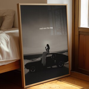 May include: A framed black and white print featuring a classic car with a person sitting on top. The image has the text "road over the ring" at the top. The print is leaning against a wall near a bed and a window.