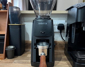 Puede incluir: Un molinillo de café Baratza negro con una tolva de plástico transparente en la parte superior y un botón de encendido central. Un portafiltro de metal con mango de madera está insertado en el soporte frontal, bajo el mecanismo de molido.