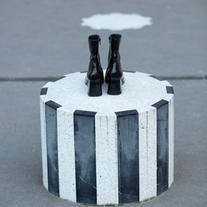 May include: A pair of black leather ankle boots with a low block heel, sitting on a round, black and white striped pedestal.