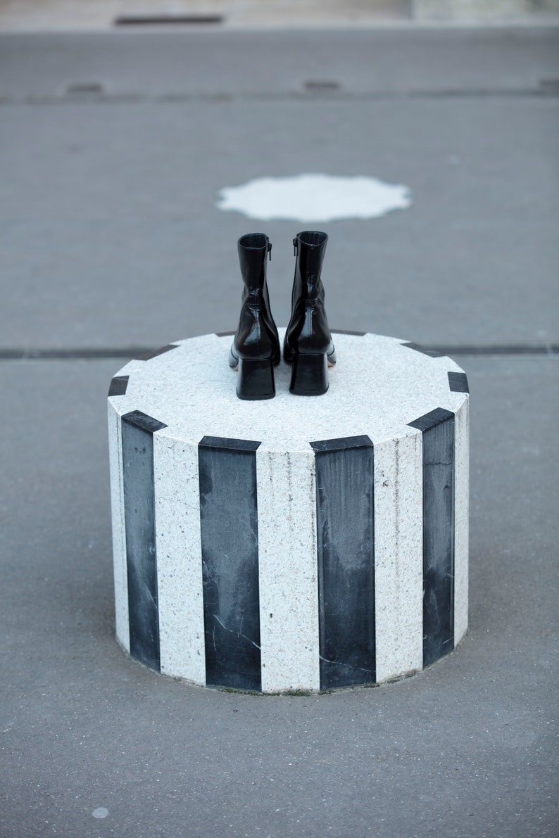 May include: A pair of black leather ankle boots with a low block heel, sitting on a round, black and white striped pedestal.
