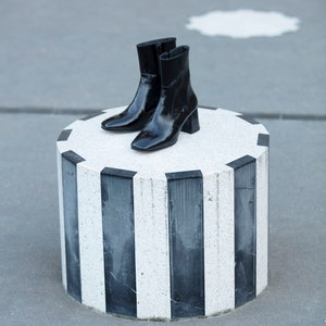 May include: A pair of black patent leather ankle boots with a block heel, sitting on a white and black striped pedestal.