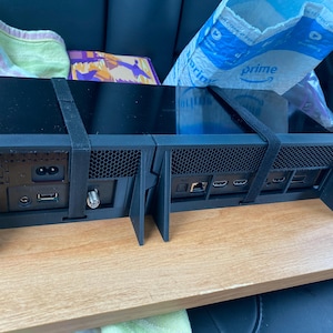 May include: Black electronic device with multiple ports and connectors on the back. The device is sitting on a wooden surface and has two black straps attached to it.