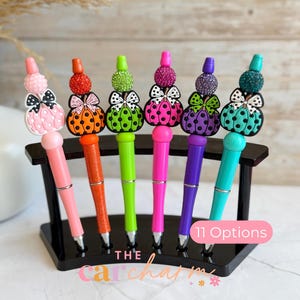 May include: Six pens arranged in a black stand, each featuring a pumpkin-shaped topper with a bow and a textured bead. The pens come in pink, orange, lime green, hot pink, purple, and teal. A pink label on the stand reads 11 Options.
