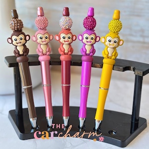 Puede incluir: Cinco bolígrafos coloridos con adornos de monos exhibidos en un soporte negro. Cada bolígrafo presenta un color diferente: marrón, rosa, rojo, morado y amarillo. Los adornos de monos están adornados con cuentas decorativas. El texto "the carcharm" es visible.