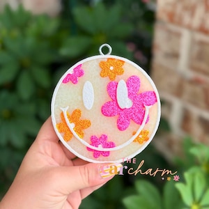 May include: A circular ornament featuring a white smiley face with glittery pink and orange flower accents. The design includes two white oval eyes and a curved white mouth. A small silver metal loop is attached to the top for hanging purposes.