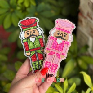 May include: Two glittery Christmas nutcracker charms. One is red, green, and white, the other is pink and white. The charms have a cartoon-like design with a hat, mustache, and uniform. The text "The Car Charm" is visible.