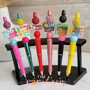 Puede incluir: Un soporte negro para bolígrafos que muestra seis bolígrafos coloridos con adornos decorativos. Los bolígrafos tienen varios diseños, incluyendo purpurina, cuentas y dijes con temas de verano con textos como "Hello Summer" y "Beach Please". La base del soporte tiene el texto "the carcharm".