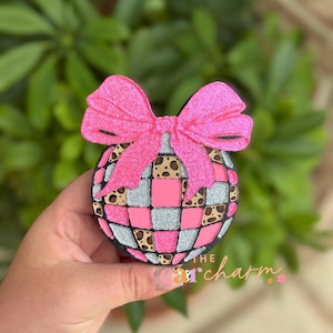 May include: A circular ornament featuring a grid pattern of pink, silver glitter, and leopard print squares. A large, bright pink glittery bow is attached to the top. The text "THE car charm" is printed in the lower right corner of the object.