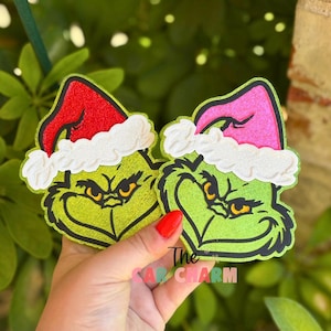 May include: Two green Grinch-themed Christmas ornaments with black outlines, wearing Santa hats with white trim. One hat is red, the other pink. The text "The Car Charm" is visible.
