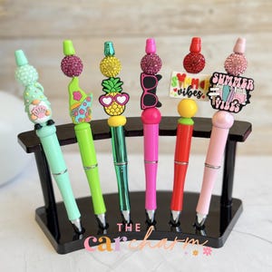 May include: Six colorful pens displayed in a black curved stand. Each pen features a unique topper including a gnome, pineapple, sunglasses, and summer-themed signs. The pens are in shades of mint, lime, green, pink, red, and light pink.