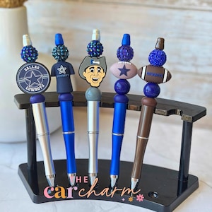 May include: Five decorative pens with blue and silver barrels, each topped with a charm. The charms feature a Dallas Cowboys logo, a jersey, a cartoon character, a football, and a beaded sphere. The pens are displayed in a black stand.