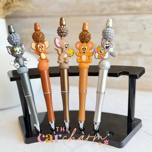 May include: Five pens featuring Tom and Jerry cartoon character toppers. The pens have silver, brown, and orange barrels, displayed on a black stand. The toppers have detailed character designs and decorative beads.