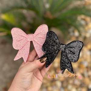 May include: Two bow-shaped ornaments, one in light pink glitter and one in black glitter, featuring a raised border design. The text "THE car charm" is printed in small, colorful letters at the bottom right of the black bow ornament.
