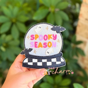 May include: A glittery snow globe ornament featuring the text SPOOKY SEASON in pink and orange letters. The design includes two white ghost figures, two black bats, and gold star accents. The base is decorated with a black and white checkerboard pattern.