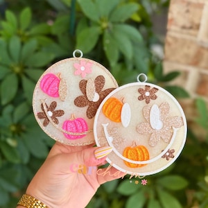 May include: Two circular ornaments featuring a smiley face design. The left ornament has pink pumpkins and brown glitter flowers on a translucent base. The right ornament has orange pumpkins and beige glitter flowers on a translucent base.