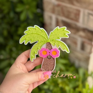 May include: A palm tree ornament featuring glittery lime green fronds and a textured brown trunk. The tree wears bright pink flower-shaped sunglasses with orange centers. The text "THE car charm" is printed in white and pink at the bottom.