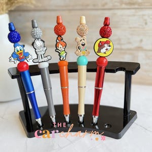 May include: A set of five decorative pens with character toppers, including Donald Duck, Garfield, and others. The pens have various colors, such as blue, gray, orange, and red, and are displayed in a black pen holder. The text "THE carcharm" is visible.