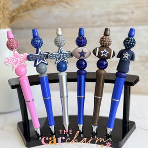Op de afbeelding: Zes pennen op een zwarte standaard, elk versierd met decoratieve kralen en bedels. De bedels bevatten sterren met de tekst COWBOYS, een football en een shirt. De pennen zijn roze, blauw, zilver en bruin van kleur met bijpassende kralen.