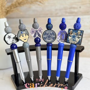 May include: Six pens featuring Dallas Cowboys themes, displayed on a black stand. The pens include charms of a football helmet, a mascot, a bow, a circular logo, a football, and a square photo. Each pen is topped with a textured, beaded accent.