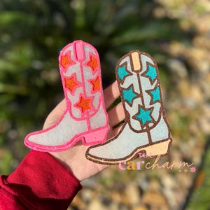 May include: Two white glitter cowboy boot car charms with different colored star designs. The boot on the left has orange stars and a pink outline. The boot on the right has teal stars and a brown outline. The text "The Car Charm" is visible in the bottom right corner of the image.