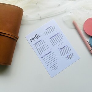 May include: A brown leather journal lies next to a printed sheet with the word "Faith" in cursive. A pink pen and two round coasters, one pink and one blue, are also on the white surface. The image has a soft, airy aesthetic.
