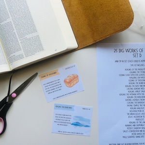 May include: An open book with a tan leather cover, two small cards, and a pair of scissors with pink accents. The cards have text and illustrations. A sheet of paper lists "25 Big Works of the Bible".