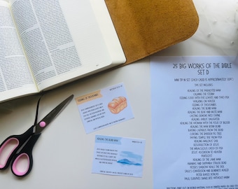 May include: An open book with a tan leather cover, two small cards, and a pair of scissors with pink accents. The cards have text and illustrations. A sheet of paper lists "25 Big Works of the Bible".