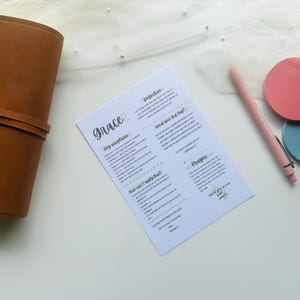 May include: A brown leather journal with a strap closure, a printed white paper with the word "Grace", a pink pen, and two round sticky notes, one pink and one blue, on a white surface.