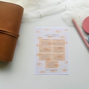 May include: A flat lay featuring a brown leather journal, a printed card with text about the 7 Feasts of Israel, a pink pen, and two round sticky notes. The card has a beige background with bread illustrations.